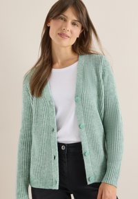 Mátová pletená vesta s knoflíkovým zapínáním, ribbovanou strukturou a kulatým výstřihem přes bílé tričko. Viditelné černé kalhoty a knoflíky.