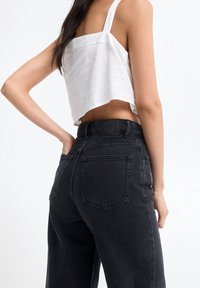 Pantalones de mezclilla negros de cintura alta con dos bolsillos traseros y una etiqueta de cuero en la cinturilla. Combinados con una blusa sin mangas blanca y corta.