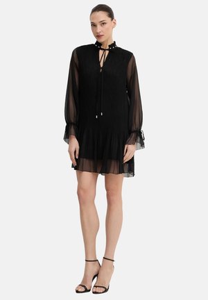 Donna che indossa un abito nero lungo in tessuto trasparente con maniche lunghe, collo e orlo arricciati, abbinato a tacchi neri con cinturino alla caviglia, in piedi su sfondo bianco.