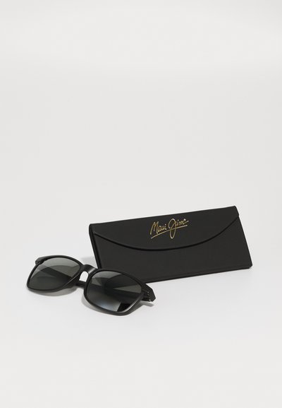 Svarte solbriller med mørke linser ved siden av et svart etui med gull "Maui Jim"-logo. Rundt rammedesign med glatt tekstur.