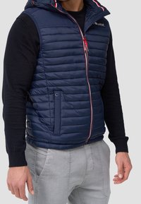 Gilet imbottito blu navy con sezioni trapuntate orizzontali, cappuccio e dettagli rossi. Caratterizzato da una cerniera e tasche laterali.