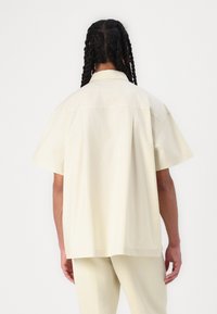 Κοντομάνικο, ανοιχτό μπεζ πουκάμισο με χαλαρή εφαρμογή, μαλακό ύφασμα και πτυχών πίσω για επιπλέον άνεση. Μινιμαλιστικός σχεδιασμός χωρίς ορατά μοτίβα.