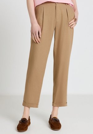 Donna che indossa pantaloni beige a vita alta con risvolti, abbinati a mocassini in camoscio marrone, in piedi su un pavimento e sfondo bianco uniforme.