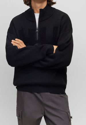 Personne les bras croisés portant un pull noir à fermeture quart zip et un pantalon cargo gris foncé sur un fond gris uni.