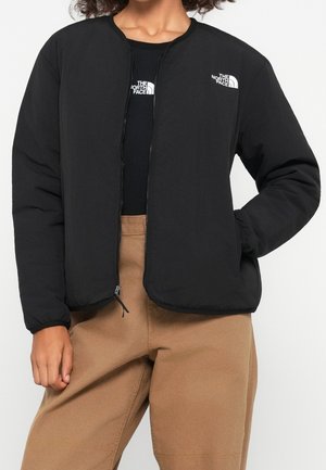 Person iført en sort North Face jakke og matchende sort top, kombineret med lysebrune afslappede bukser, stående mod en ensfarvet baggrund.