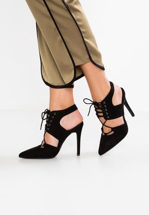 Nogi w beżowych spodniach z czarnym obszyciem oraz czarne sznurowane buty na wysokim obcasie z wycięciami po bokach na białej podłodze.