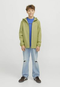 Ljusgrön, vattenresistent jacka med huva, framsida med dragkedja och elastiska ärmmuddar, lager över en blå tröja och i kombination med slitna jeans.