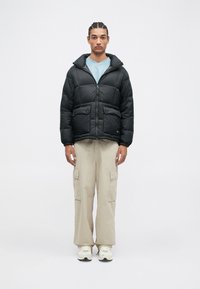 Μαύρο επενδύστηκες μπουφάν με φερμουάρ μπροστά, που διαθέτει δύο μεγάλες τσέπες και κουκούλα, φορεμένο πάνω από ένα ανοιχτό μπλε πουκάμισο και συνδυασμένο με μπεζ παντελόνι cargo.