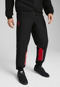 Pantalons de survêtement noirs avec des accents rouges sur les côtés, taille élastique et chevilles resserrées. Présente un logo sur la cuisse. Associés à des baskets noires.