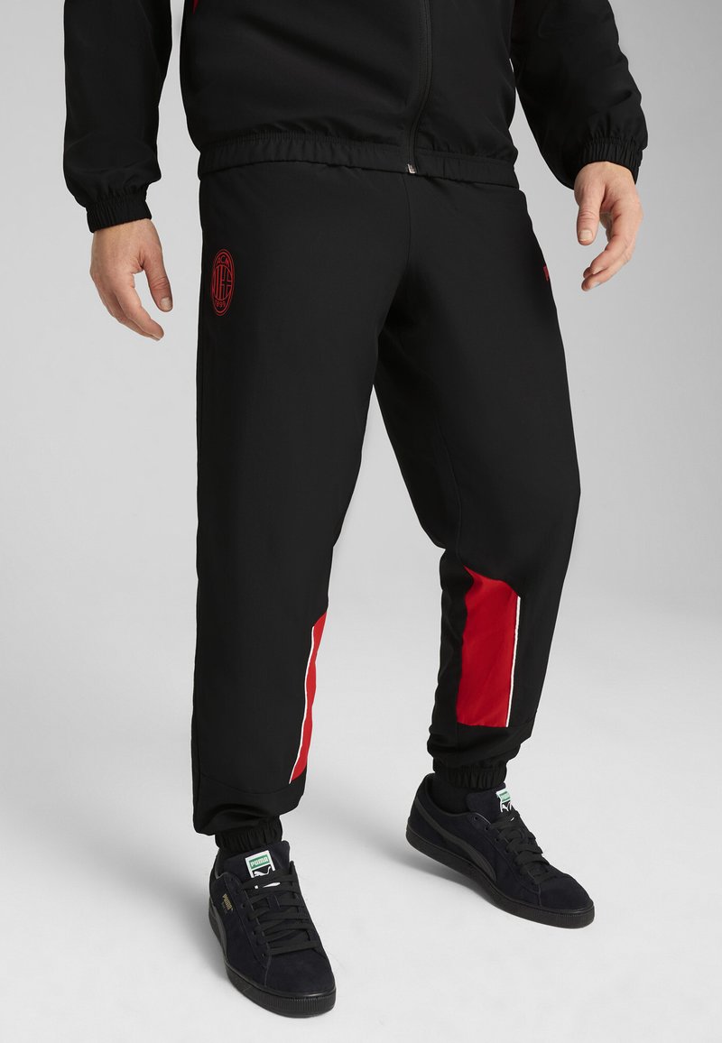 Schwarze Jogginghose mit roten Seitenakzenten, elastischem Bund und Bündchen an den Knöcheln. Mit einem Logo am Oberschenkel. Kombiniert mit schwarzen Turnschuhen.