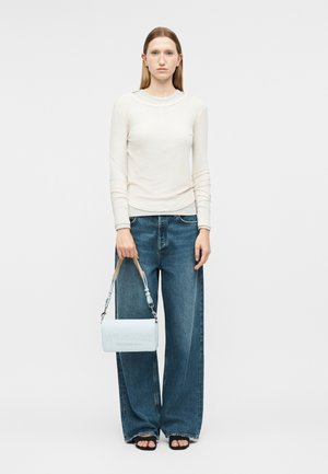 Jonge vrouw in een wit shirt met lange mouwen en wijd uitlopende jeans, die een witte Love Moschino handtas met een beige bandje vasthoudt.