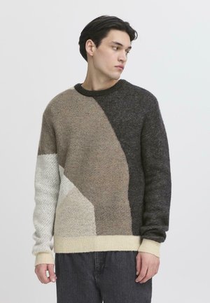 Young man wearing a crewneck sweater with beige, dark gray, and cream abstract color blocks, looking to his left against a plain background.