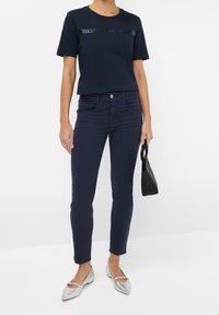 Námořnické tričko s flitrovým detailem na hrudi, v kombinaci s námořnickými slim-fit džíny. Modelka drží černou kabelku a nosí stříbrné baleríny.