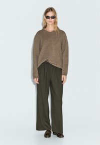 Maglione marrone di grandi dimensioni a maglia con scollo a V, abbinato a pantaloni larghi a gamba ampia di un verde scuro. Gli accessori includono occhiali da sole scuri e scarpe marroni.