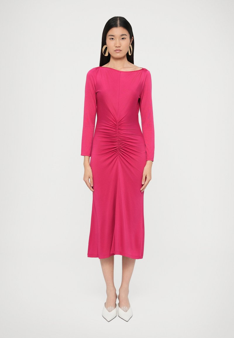 Fukszia hosszú ujjú ruha, deréknál gyűjtött részletekkel, áramló sziluettel, sima textúrával és hajónyak kivágással. Fehér hegyes cipővel párosítva.