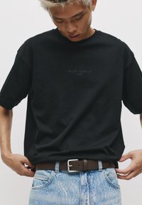 T-shirt en coton noir avec un col rond et des manches courtes, avec un texte brodé "JE NE ME SUIS PAS RÉVEILLÉ (ENCORE)" sur le devant. Porté avec un jean bleu clair.