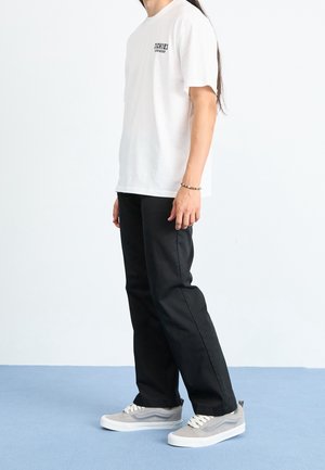 Vit bomullströja med svart logotyp på bröstet, svarta byxor och grå sneakers med vita sulor och dekorativ sömnad på sidorna.