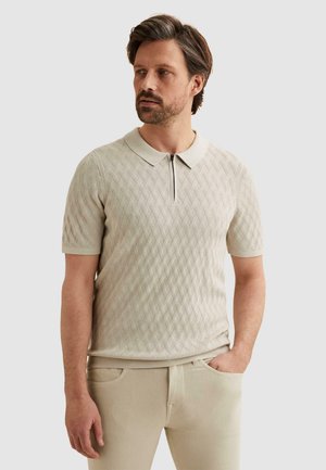 Mann mit kurzem Bart, der ein beiges, kurzärmeliges, strukturiertes Poloshirt mit Reißverschluss und passende Hose trägt, steht mit einer Hand in der Tasche.