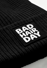Black knitted beanie with a textured pattern. Features a rectangular, embroidered patch displaying "BAD HAiR DAY" in bold white letters.