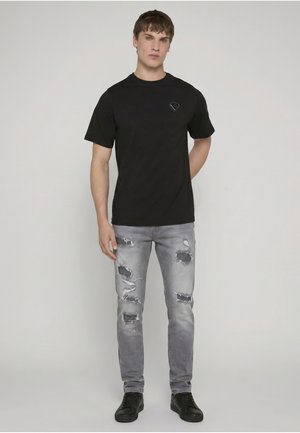 Młody mężczyzna stojący, ubrany w czarny t-shirt z krótkim rękawem i małym logo na piersi, podarte szare dżinsy oraz czarne trampki, neutralne tło.