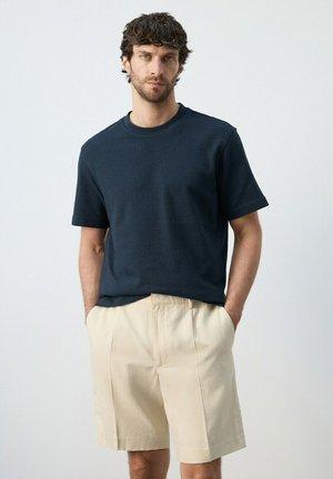 Hombre con cabello rizado y oscuro, vistiendo una camisa azul marino de manga corta y pantalones cortos beige hasta la rodilla, de pie con las manos en los bolsillos.