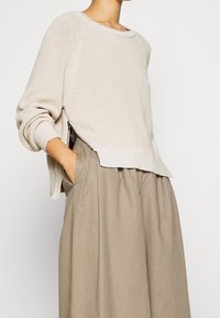 Pull beige clair en maille avec une coupe décontractée, des poignets côtelés et des fentes sur les côtés. Associé à une jupe fluide de couleur taupe.