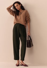 Pulover cămilă cu nasturi, pantaloni croiți verde închis, pantofi balerini marsala și o geantă mică maro. Texturi moi, croială lejeră și linii clare.