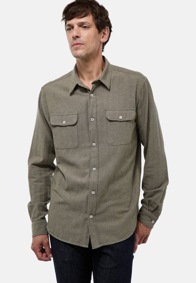 Chemise à manches longues en tissu vert olive, avec un col button-down, deux poches poitrine et des accents de boutons blancs. Texture lisse.