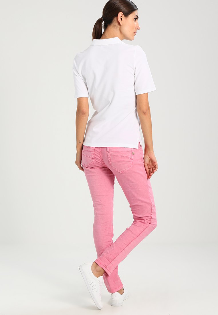 Frau mit braunen Haaren, die nach hinten gebunden sind, trägt ein weißes Polohemd mit kurzen Ärmeln, rosa Skinny-Jeans und weiße Sneakers und steht mit gekreuzten Beinen.