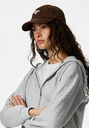 Femme aux longs cheveux bouclés et foncés, portant une casquette de baseball marron, un sweat à capuche gris zippé et un t-shirt blanc, les bras croisés, regardant sur le côté.