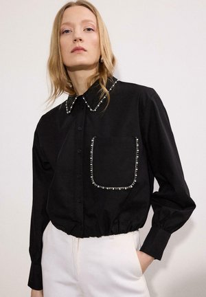 Plavovlasá žena v čiernej košeli s gombíkmi a perlovou ozdobou na golieri a vrecku, v kombinácii s bielymi nohavicami, stojaca pred jednoduchým pozadím.