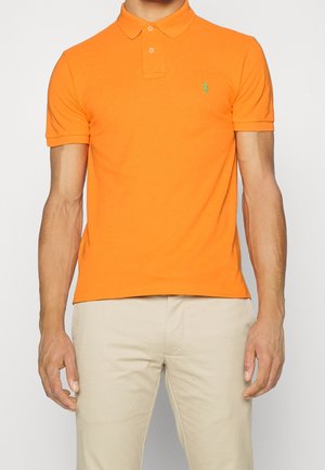 Mężczyzna w krótkim rękawie jaskrawo pomarańczowego polo z małym zielonym logo i jasnobeżowych spodniach, stojący na tle jednolitego tła.