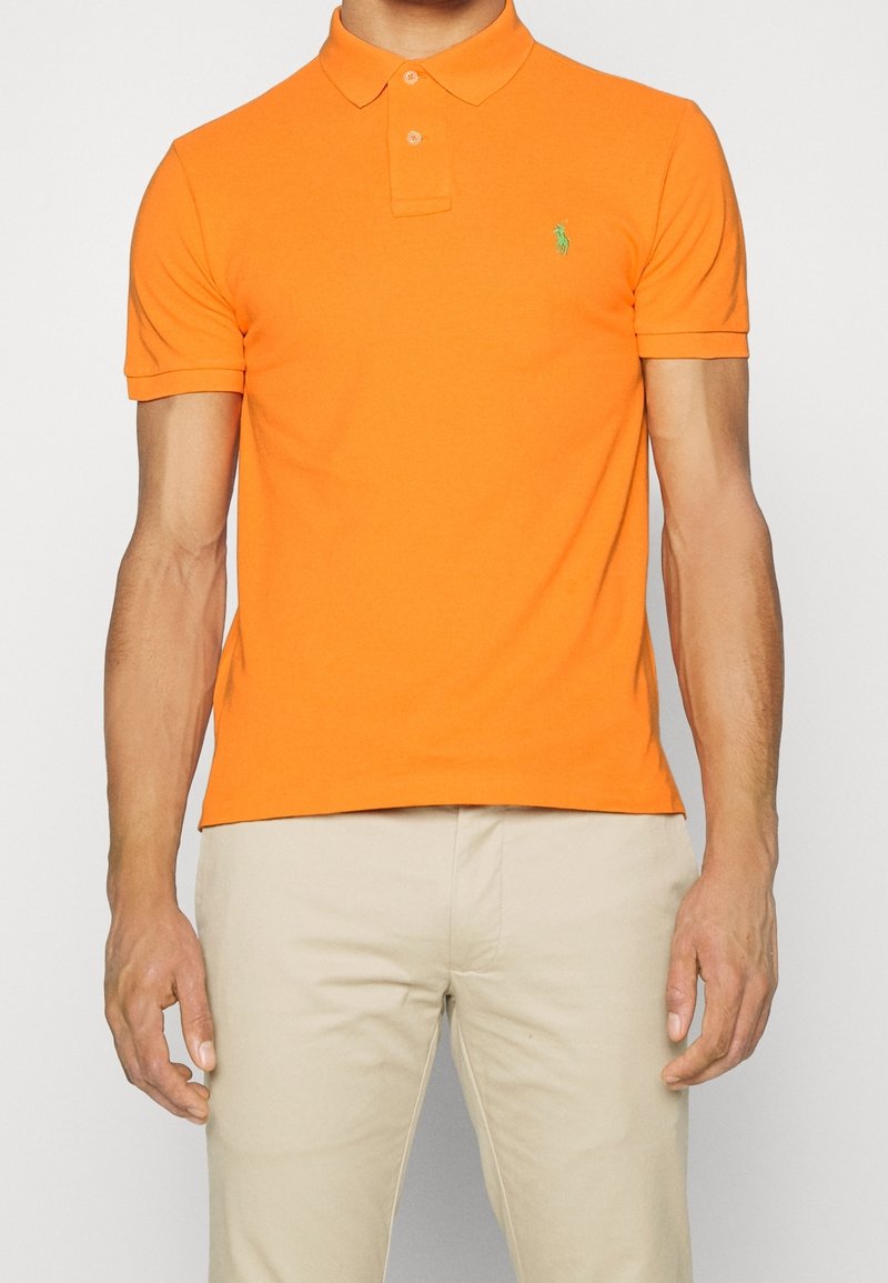 Homme portant un polo à manches courtes orange vif avec un petit logo vert et un pantalon beige clair, debout devant un fond uni.