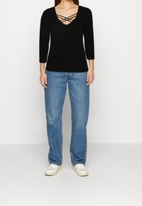 Schwarzes Langarmoberteil mit V-Ausschnitt und gekreuzten Trägerdetails, kombiniert mit hellblauen gerade geschnittenen Jeans und weißen Sneakers.
