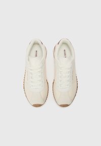 Sneakers bianchi con lacci, dalla parte superiore texturizzata e dettagli in beige chiaro. Presentano una suola in gomma con un motivo a zigzag e un brand discreto.