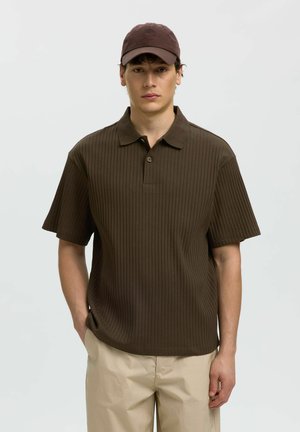 Junger Mann, der ein braunes, geripptes kurzärmliges Polohemd, beigefarbene Hosen und eine passende braune Baseballkappe trägt, steht mit einer Hand in der Tasche.