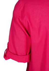 Manche de chemise en lin rose vif roulée avec boucle en tissu et détail de bouton sur fond blanc.