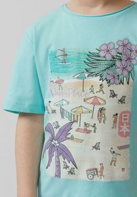 Lichtblauw katoenen t-shirt met een kleurrijke strandafbeelding, inclusief parasols, palmbomen en mensen die verschillende activiteiten ondernemen.