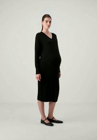 Μαύρο πλεκτό μίνι φόρεμα εγκυμοσύνης, με V-λαιμόκοψη, μακριά μανίκια, ribbed υφή, μήκους μέχρι το γόνατο. Συνδυασμένο με μαύρες μπαλαρίνες, το μοντέλο στέκεται μπροστά από έναν ουδέτερο φόντο.