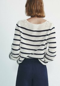 Vrouw met de rug naar de camera, gekleed in een wit en marineblauw gestreepte gebreide trui en een marineblauwe broek, tegen een effen lichte achtergrond.