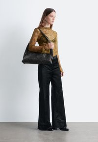 Schwarze geflochtene Tragetasche mit strukturierter Oberfläche, kombiniert mit einem transparenten, olivgrünen Langarmoberteil und weiten, schwarzen Samthosen.