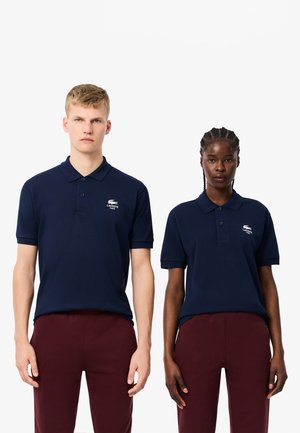 Polo navy in cotone, a maniche corte, con colletto piatto e un piccolo logo ricamato sul petto. Indossati con pantaloni bordeaux.