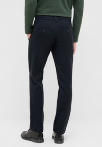 Marineblaue Hose mit geradem Schnitt und sichtbaren Gesäßtaschen, gefertigt aus glattem Material. Kombiniert mit schwarzen Schuhen und einem grünen Oberteil.