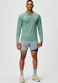 Langärmliges grünes Sportshirt mit dunklem Logo, kombiniert mit hellgrauen Shorts mit marineblauen Akzenten. Getragen mit grauen Socken und Schuhen.