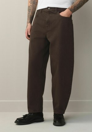 Mann trägt dunkelbraune Hose mit geradem Bein, schwarze Leder-Tassel-Loafers und ein weißes Hemd, sichtbar sind tätowierte Arme.