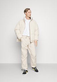 Beige pufferjakke med høj krave, parret med lysebeige bukser. Sorte sneakers med gule striber. Minimalistisk design, bløde teksturer.