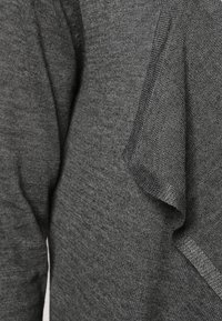 Zbliżenie na ciemnoszary, dziany materiał ukazujący teksturowane, opadające fałdy oraz długie rękawy na odzieży.