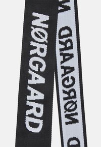 Černo-bílý tkaný popruh s tučným textem. Popruh má plochý, texturovaný design s kontrastními barevnými nitěmi, které tvoří slovo "Nørgaard."