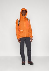 Lys orange, let jakke med hætte, der har lynlåslommer og kontrasterende grå bukser. Sorte vandresko med farverige detaljer.