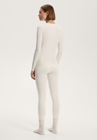 Weißes Langarmoberteil und Leggings-Set, körperbetontes Design mit glatter Textur. Das Modell trägt gerippte Socken und ist von hinten vor einem neutralen Hintergrund zu sehen.